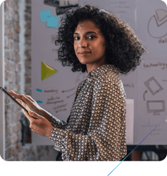 A woman with dark curly hair is shown in a professional setting, looking directly at the camera while holding a tablet in her hands. Behind her is a whiteboard with some diagrams and text.